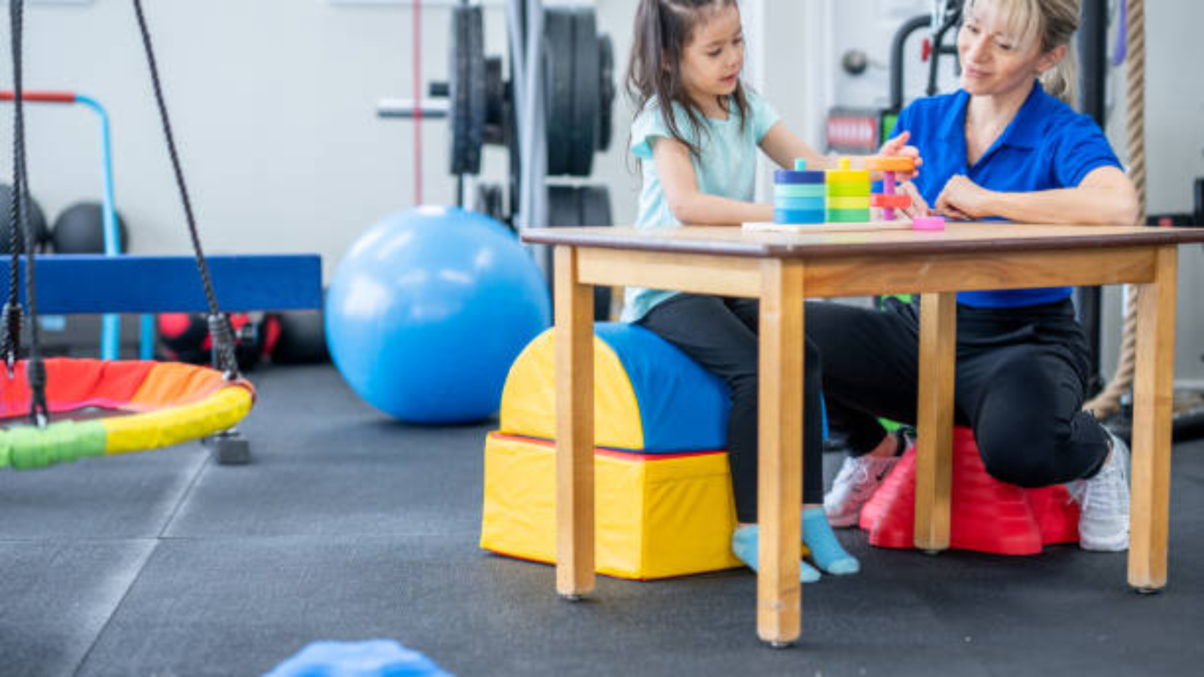A female therapist sits with a little girl at a table as they work on her motor skills.  The therapist is dressed professionally and is using colorful toys to help her build strength in her hands and coordination.